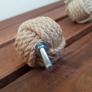 May include: A close-up of a brown rope knob with a silver screw. The knob is made of natural rope and has a knot design. It is a decorative hardware piece for furniture or cabinets.