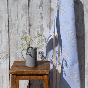 May include: A light blue apron with a white fern pattern hangs on a white wood wall. A grey ceramic jug with white flowers sits on a wooden stool.