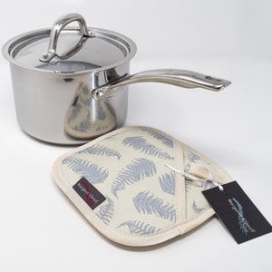 May include: A silver saucepan with a lid and a handle. The saucepan is sitting on a white pot holder with a fern pattern. The pot holder has a tag that says "Margaret Clavell".
