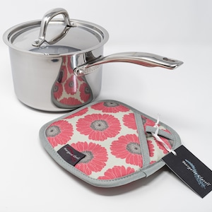 May include: A stainless steel saucepan with a lid and a silver handle. The saucepan is sitting on a pink and white floral pot holder with a grey border. A black tag with white text reads "Margaret & Maxwell"