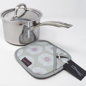 May include: A silver saucepan with a lid and a matching handle. The saucepan is sitting on a grey and white floral pot holder with a red tag that says "Margaret Russell".