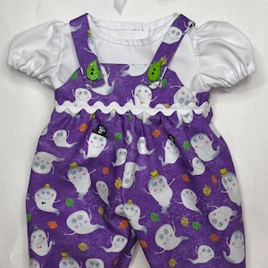 May include: A purple overall outfit with a white short-sleeved shirt featuring a ghost and autumn leaf print. The overalls have white rickrack trim, green leaf-shaped buttons on the straps, and a matching headband with a white ribbon bow.