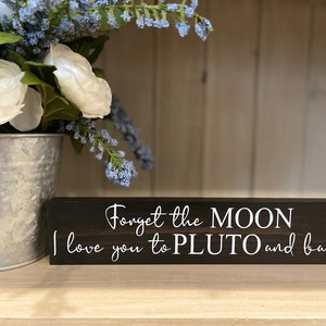 以下が含まれることがあります： 「Forget the MOON I love you to PLUTO and back」と書かれた黒い木製の看板です。