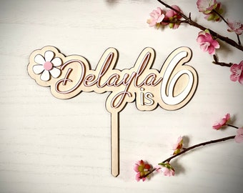 Può includere: Topper per torta in legno con il nome "Delayla" e il numero "6" in scrittura rosa. Il topper include anche un fiore bianco con un centro rosa e le parole "is 6" in bianco.