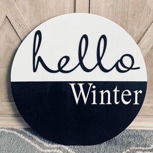 May include: A round, black and white wooden sign with the text "hello winter" in white lettering on a black background and "hello" in black lettering on a white background.