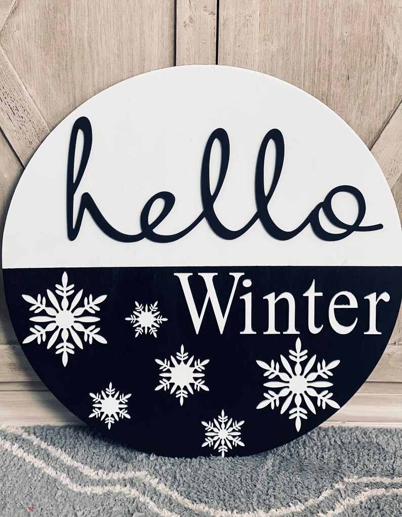 May include: A black and white round wooden sign with the text "hello winter" in black lettering. The sign is decorated with white snowflakes.