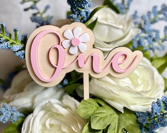 May include: Pink wooden "One" cake topper with a white flower and pink center. The topper is surrounded by white and blue flowers.