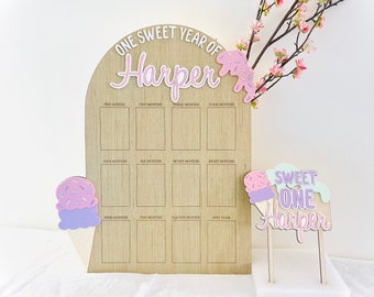 Op de afbeelding: Een houten groeikalender met een roze en wit ijshoorntjesontwerp. De kalender is gelabeld "One Sweet Year of Harper" en heeft ruimte voor elke maand van het jaar.