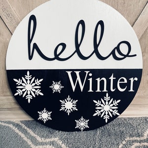 May include: A round wooden sign with a black and white design. The top half is white with the word "hello" in black cursive lettering. The bottom half is black with the word "Winter" in white lettering and white snowflake designs.