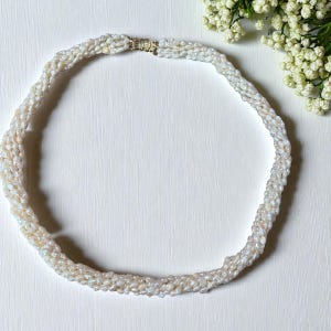 Puede incluir: Un collar de perlas blancas con un diseño trenzado. El collar está hecho de pequeñas perlas irregulares y tiene un cierre dorado. El collar se muestra sobre una superficie blanca, con flores blancas en el fondo.
