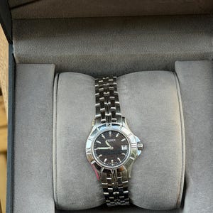 Op de afbeelding: Een zilveren Fendi polshorloge met een zwarte wijzerplaat, datumweergave en zilveren wijzers en markeringen. Het horloge heeft een roestvrijstalen band en wordt gepresenteerd in een grijze horlogedoos.