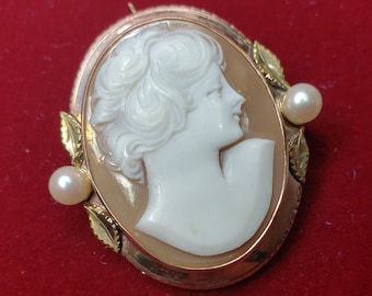 Puede incluir: Un broche de camafeo dorado con un camafeo blanco de perfil de mujer. El camafeo está rodeado por un marco dorado con dos pequeños acentos de perla.