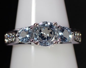 May include: A silver ring with three blue gemstones set in a row. The ring is on a white display stand.