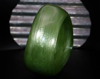 May include: A smooth, olive green bangle bracelet. The bracelet has a rounded, oval shape and a glossy finish. The background is dark with a textured, silver-colored element. The bracelet is a piece of jewelry.