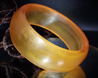 May include: A translucent, amber-colored bangle bracelet. The smooth, rounded bracelet has a warm, golden hue with subtle variations in color. The light catches the surface, creating highlights and shadows. The jewelry is a simple, elegant accessory.