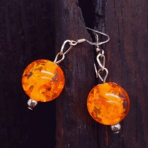 May include: A pair of orange amber earrings with silver hooks. The earrings are round and have a marbled pattern.