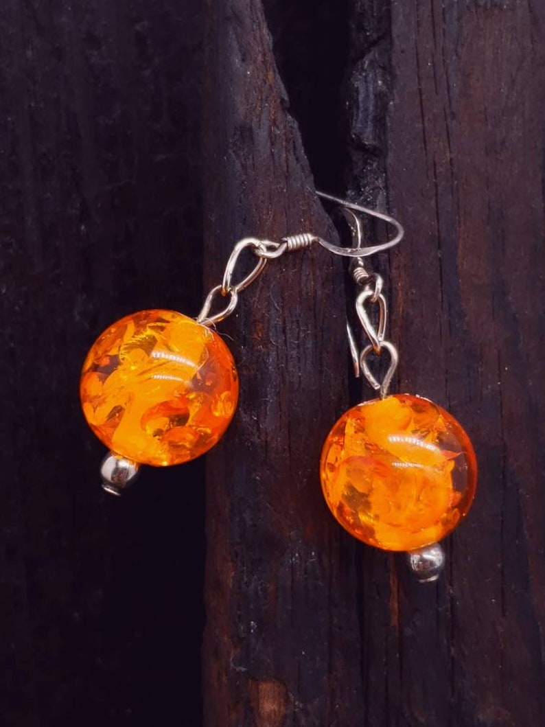 May include: A pair of orange amber earrings with silver hooks. The earrings are round and have a marbled pattern.