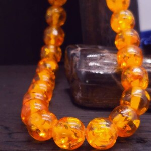 May include: A close-up of a necklace made of amber beads. The beads are a deep orange colour and are strung together on a clear string. The necklace is lying on a dark wooden surface.