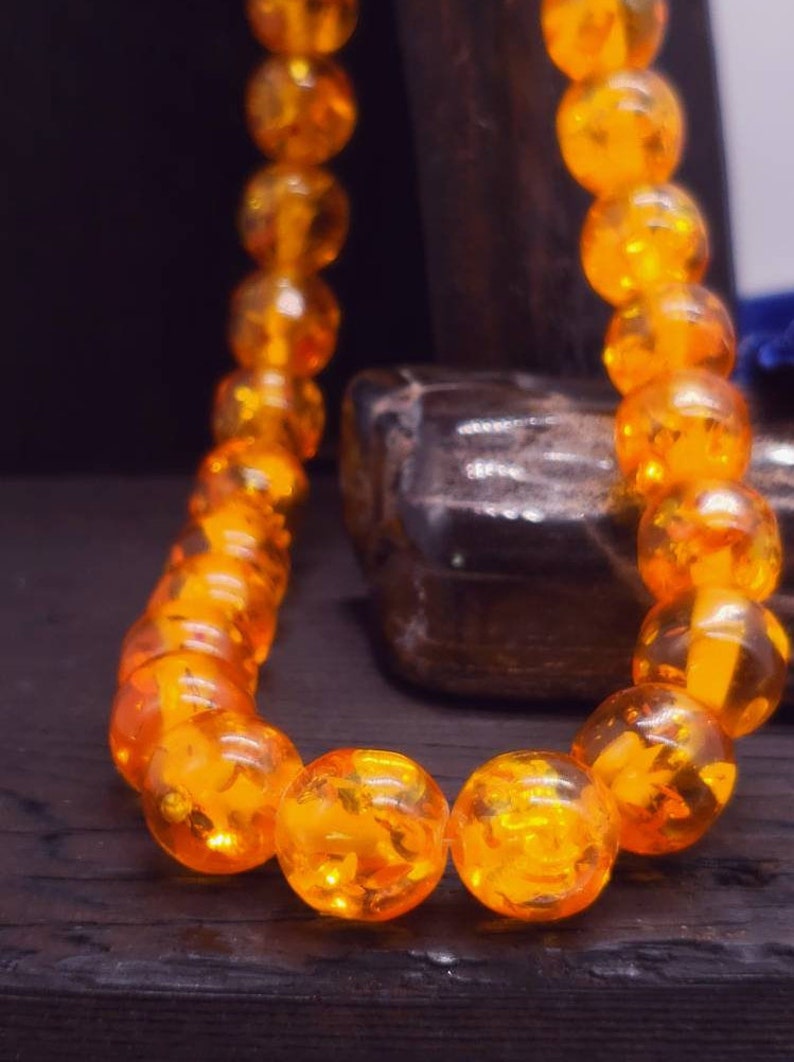 May include: A close-up of a necklace made of amber beads. The beads are a deep orange colour and are strung together on a clear string. The necklace is lying on a dark wooden surface.