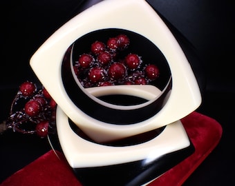 May include: Two black and white plastic bangle bracelets with a square shape. The bracelets are stacked on top of each other.