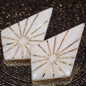 Könnte beinhalten: Ein Paar weiße, rautenförmige Ohrringe mit goldenen Akzenten. Die Ohrringe haben ein Sonnenstrahl-Design mit goldenen Linien, die vom Zentrum ausstrahlen.