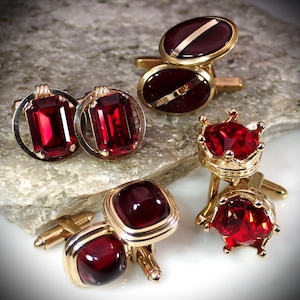 May include: An assortment of gold-toned cufflinks with various designs. The cufflinks feature vibrant red gemstones in oval, rectangular, and crown shapes. Some have gold accents, creating a luxurious and classic aesthetic.