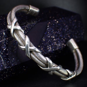 Può includere: Bracciale a polsino color argento con un design intrecciato e incrociato. Il bracciale presenta un aspetto strutturato e intrecciato e una finitura lucida. Il design incorpora fili spessi e sottili, creando un motivo dettagliato e complesso.
