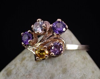 May include: A gold ring set with five gemstones. The ring has a delicate, swirling design with a yellow, white, and three purple gemstones.