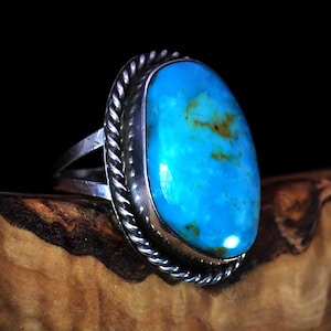 May include: A silver ring with a large, oval turquoise stone. The stone is a vibrant blue with brown and yellow inclusions. The ring has a twisted rope design around the stone and a split band.