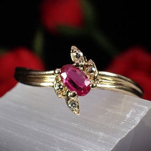 May include: A gold ring featuring an oval ruby gemstone at its centre. The ring has a swirling design with small diamond accents. It is set against a white surface with a blurred red and black background.