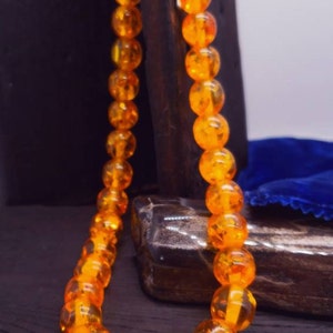 May include: A necklace made of amber beads. The beads are a deep orange colour and have a translucent quality. The necklace is strung on a silver chain.
