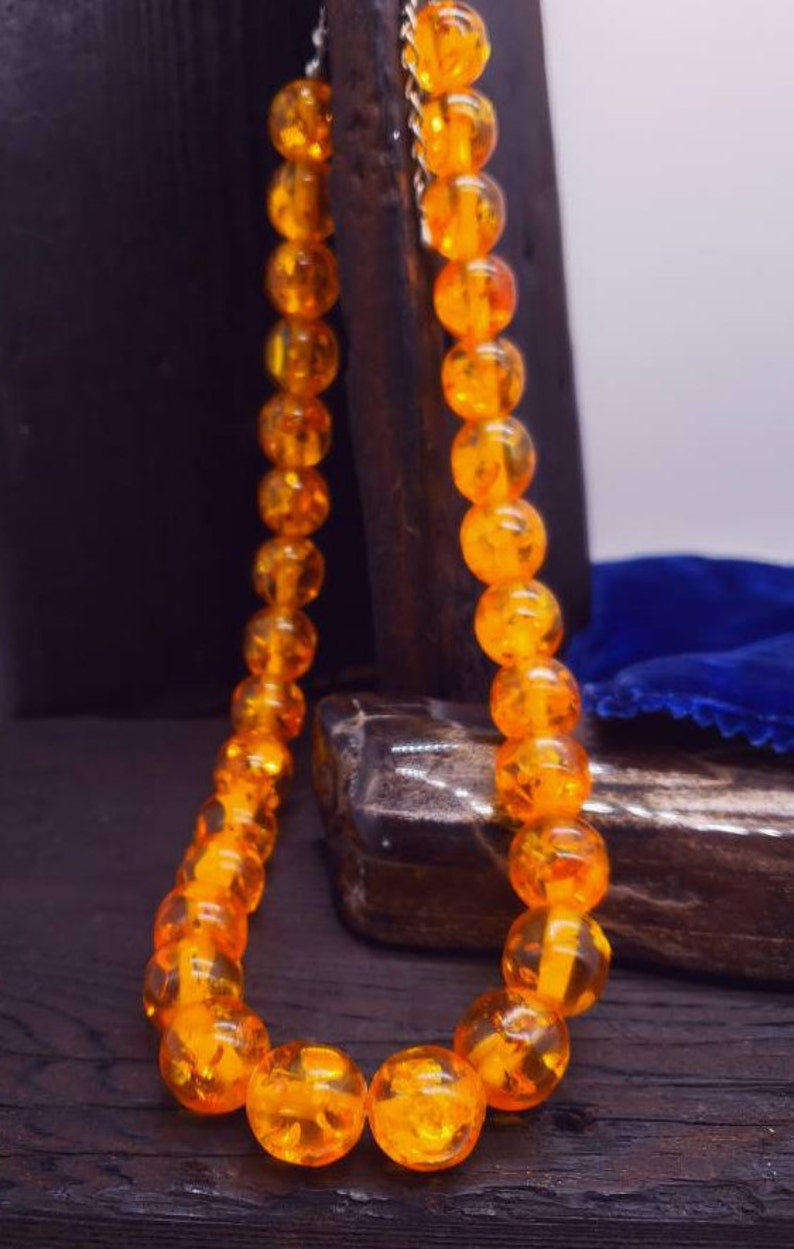 May include: A necklace made of amber beads. The beads are a deep orange colour and have a translucent quality. The necklace is strung on a silver chain.