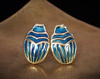 May include: A pair of gold-toned earrings shaped like scarab beetles. The beetles are blue with gold accents and have a detailed design.