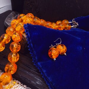 May include: A necklace and earrings set made of amber beads. The necklace is made of round, orange amber beads and the earrings are made of two round, orange amber beads each. The set is displayed on a blue velvet pouch.
