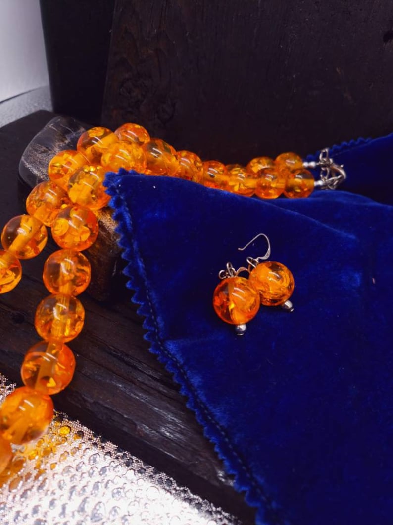 May include: A necklace and earrings set made of amber beads. The necklace is made of round, orange amber beads and the earrings are made of two round, orange amber beads each. The set is displayed on a blue velvet pouch.