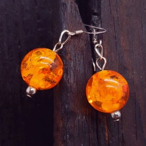 May include: A pair of orange amber earrings with silver hooks. The earrings are round and have a textured surface.