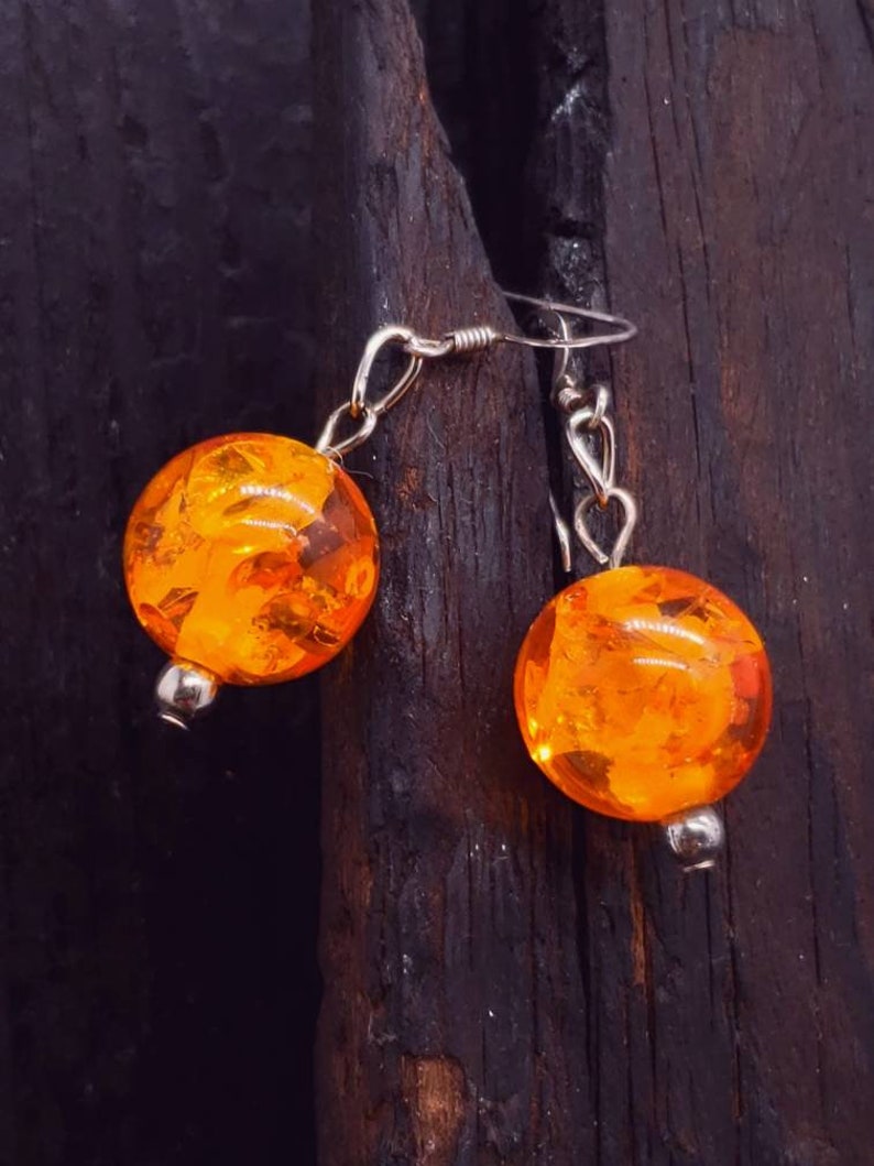 May include: A pair of orange amber earrings with silver hooks. The earrings are round and have a textured surface.