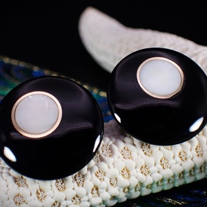 Puede incluir: Un par de pendientes redondos negros y blancos con un borde dorado. Los pendientes tienen un centro blanco y un anillo exterior negro.