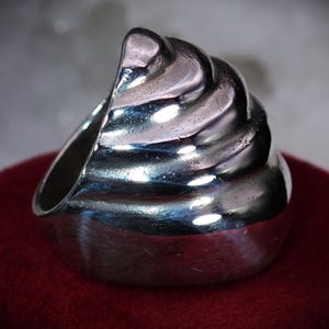 May include: A silver-colored ring with a unique design. The ring features a swirling, textured top, resembling a soft-serve ice cream cone. The ring is set on a red surface.