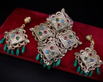 Puede incluir: Un conjunto de joyas vintage en tono dorado que incluye un broche y dos pares de pendientes. Cada pieza tiene un diseño cuadrado con piedras preciosas rojas, verdes y azules. Los pendientes tienen colgantes verdes en forma de lágrima. El conjunto se exhibe sobre una superficie de terciopelo rojo.