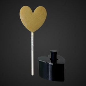 May include: A golden heart-shaped object on a white stick, and a black heart-shaped base. The text "Winay's crafts" is printed on the stick and the base. The background is a dark gray.