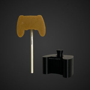 Puede incluir: Una piruleta marrón en forma de mando de juego en un palo blanco. La piruleta está hecha en forma de mando de videojuego. La piruleta está sentada en un molde de plástico negro con el texto "Winay's Crafts" en él.