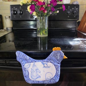 May include: A chicken-shaped fabric oven mitt featuring a blue and white paisley pattern on the body and a white patch with a blue line-art illustration of chickens. The mitt has a small orange fabric comb and beak detail at the top.
