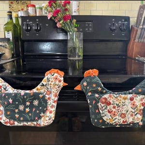 May include: Two chicken-shaped oven mitts. One mitt is white with a floral pattern, and the other is dark green with a floral pattern. Both have orange comb and beak details. Kitchen decor.