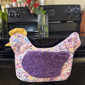 May include: A fabric chicken-shaped oven mitt featuring a white base with a colorful floral pattern of small purple, blue, and yellow flowers. It has a solid yellow fabric comb on top and a dark purple wing patch with a black paisley print design.