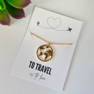Puede incluir: Un collar de oro con un colgante de mapa del mundo. El collar está en una tarjeta blanca con el texto "TO TRAVEL is to live".