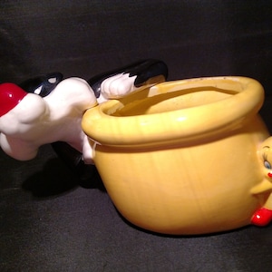 May include: A yellow ceramic pot shaped like a bag with a black and white cartoon character peeking out of the top. A yellow cartoon bird with red shoes is on the side of the pot.