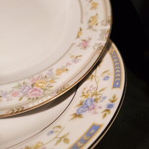 May include: Two white ceramic plates with floral patterns. The top plate has a gold trim and a delicate floral design in shades of pink, yellow, and blue. The bottom plate has a blue trim and a larger floral design with blue, pink, and yellow flowers.