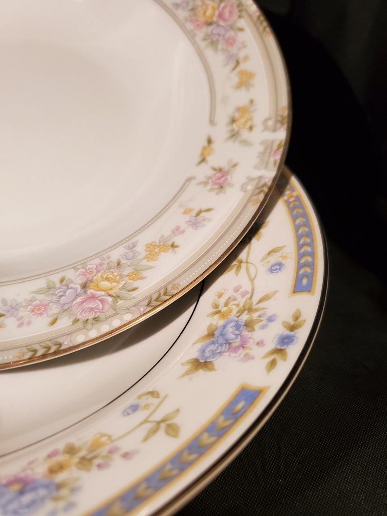 May include: Two white ceramic plates with floral patterns. The top plate has a gold trim and a delicate floral design in shades of pink, yellow, and blue. The bottom plate has a blue trim and a larger floral design with blue, pink, and yellow flowers.
