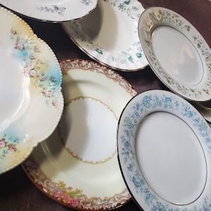 Może przedstawiać: Kolekcja siedmiu okrągłych talerzy ceramicznych z różnymi wzorami dekoracyjnymi. Talerze przedstawiają motywy kwiatowe w odcieniach różu, błękitu i zieleni, niektóre ze złotymi obrzeżami i falistymi krawędziami, ułożone w nakładającą się grupę.